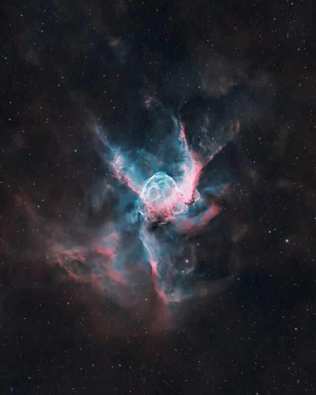 A vibrant nebula glows in deep space, with swirling clouds of blue and pink gas against a dark, star-speckled background. The nebula appears luminous at its center, radiating wisps outward.
