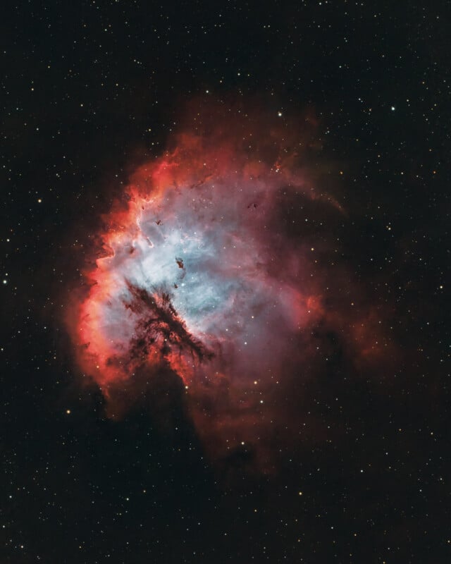 A bright, colorful nebula in deep space glows with vivid red, pink, and blue hues, surrounded by dark dust clouds and scattered stars against a black background.