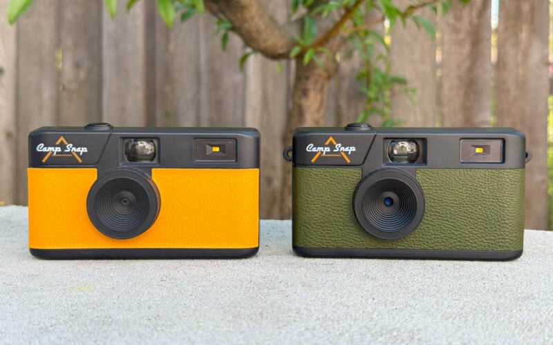 Two vintage-style cameras are displayed on a table against a wooden fence and foliage backdrop. One camera is orange and black, while the other is green and black. Both have a "Camp Snap" logo and a simple lens design.