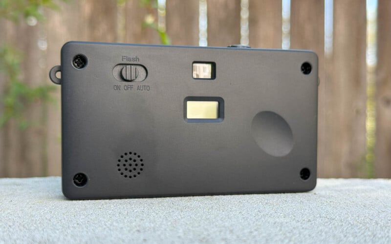 A compact black camera is shown on a surface, with visible features including a flash switch labeled "ON OFF AUTO," a flash bulb, a viewfinder, and a small speaker. A wooden fence is blurred in the background.