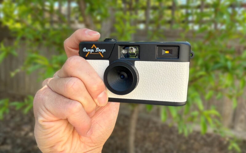 A close-up of a hand holding a compact camera with a black and white design. The camera has "Camp Snap" written on it. Blurred greenery is visible in the background.