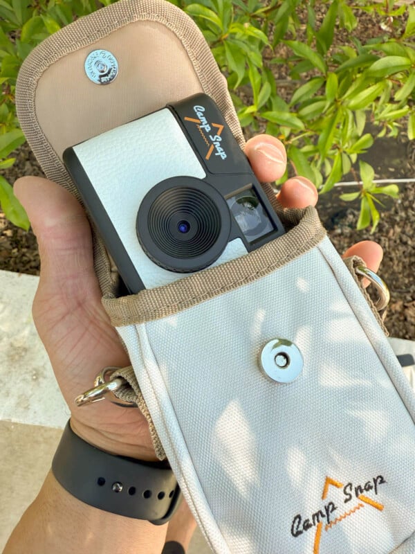 A person holds a small, vintage-style camera partially out of a beige fabric pouch with "Camp Snap" text. The background features green leaves. The person is wearing a black wristband.