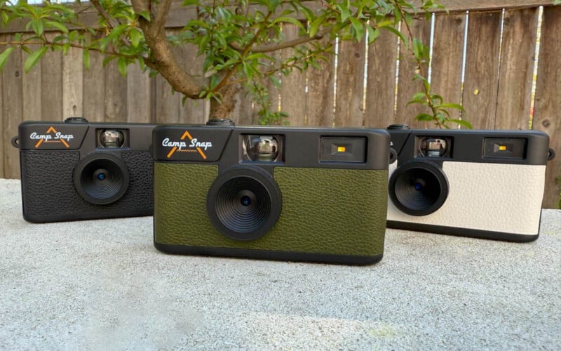 Three vintage-style cameras are displayed outdoors on a concrete surface. They have textured fronts in black, green, and white. A wooden fence and leafy tree are in the background.