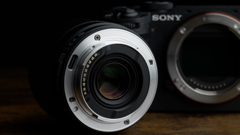 Close-up of a camera lens facing a Sony camera body with no lens attached, placed on a dark wooden surface. The camera and lens are partially illuminated, highlighting the metallic mount and electronic contacts.