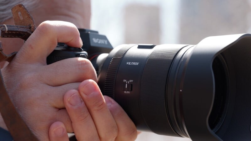 Close-up of hands holding a camera with a Viltrox lens. The camera is being adjusted for focus, and a blurred cityscape is visible in the background.