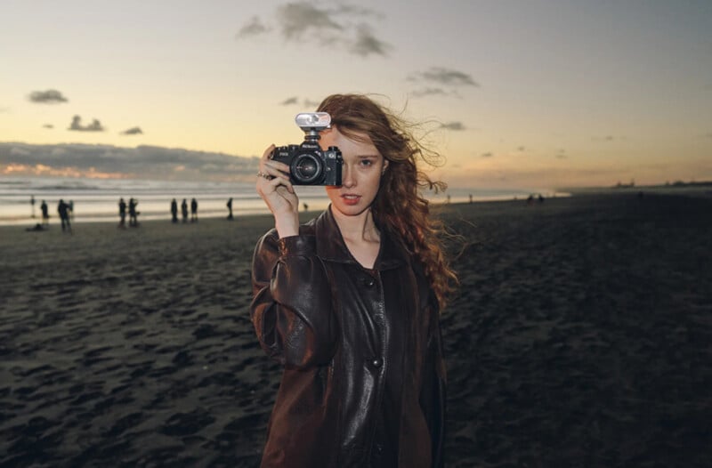 A woman with long, wavy hair stands on a beach at sunset, holding a camera up to her face. Other people are visible in the background, walking along the sand near the shoreline.