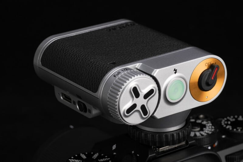 A close-up of a modern camera accessory with a textured black grip, silver dial labeled "FULL/1/2," gold on/off switch, green button, and ports, mounted on a camera body against a black background.