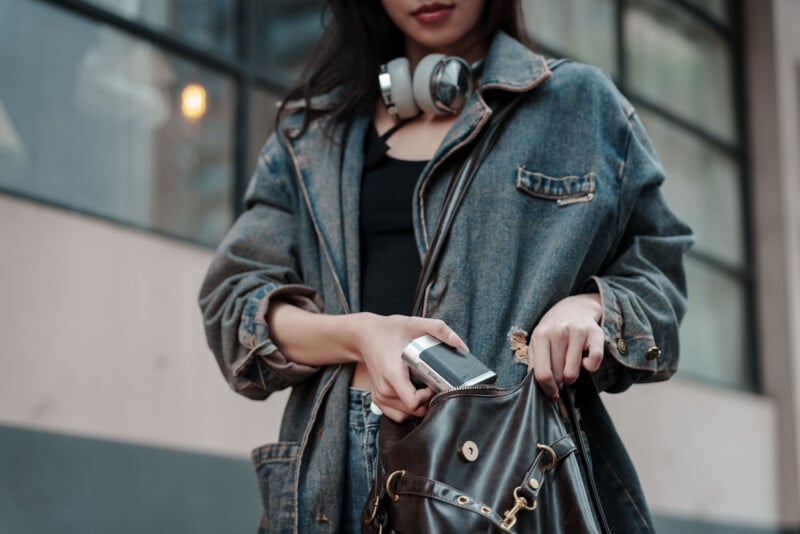 A woman wearing a denim jacket and headphones around her neck is putting a smartphone into a black leather bag while standing outside near a building with large windows.