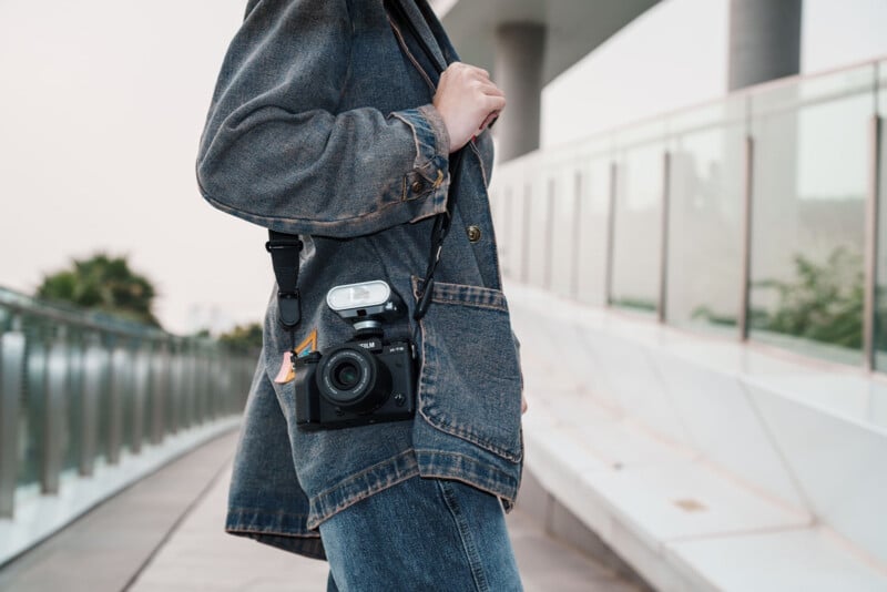 A person wearing a denim jacket and jeans stands on a modern walkway, holding a camera with a flash attached, slung over their shoulder by a strap. The background features glass railings and blurred outdoor scenery.
