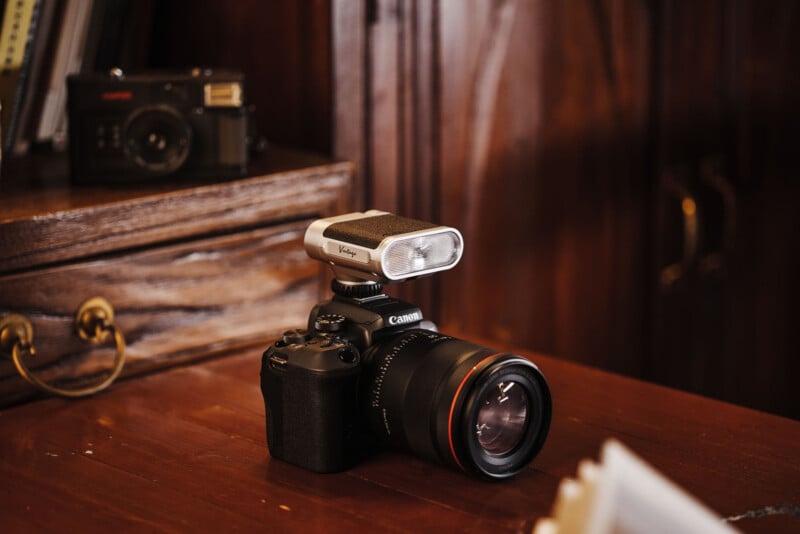 A Canon camera with a large lens and an external microphone mounted on top sits on a wooden desk. In the background, an old film camera rests on a wooden drawer.