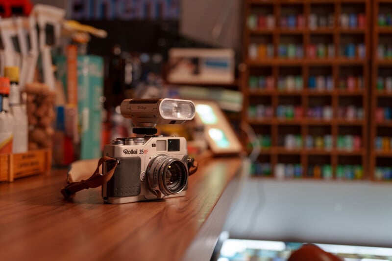 A vintage Rollei 35 S camera with a flash attachment sits on a wooden countertop in a warmly lit shop, with shelves of colorful items blurred in the background.