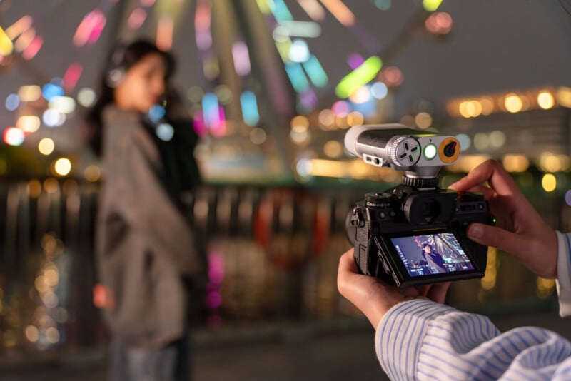 A person films a woman standing in front of a brightly lit Ferris wheel at night. The camera’s screen displays the woman and the colorful lights in the background, with the scene slightly blurred.