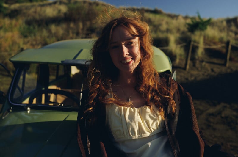 A woman with long, wavy red hair smiles at the camera, standing outdoors in front of a vintage green car. Sunlight highlights her hair, and grassy hills are visible in the background.