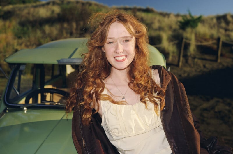 A young woman with long, curly red hair smiles outdoors, wearing a cream top and brown jacket. She stands in front of a vintage green car, with grassy hills in the background under a clear sky.