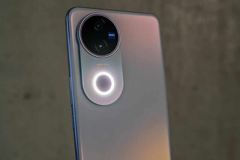 Close-up of a smartphone's rear camera module, featuring a large circular design with two lenses and a small text that says "Vivo ZEISS." The phone has a sleek, metallic finish, and the background is a blurred gray surface.