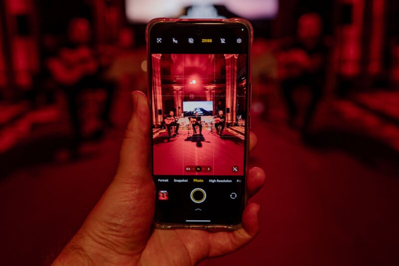 Close-up of a hand holding a smartphone, capturing a photo of musicians performing under red stage lights. The phone screen displays the band and instruments in focus, contrasting with the dark, blurred background.