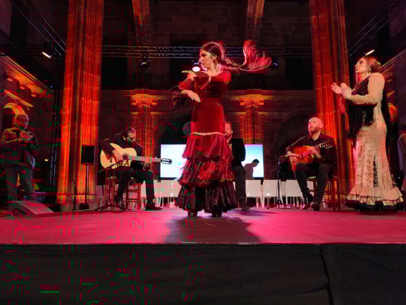 A flamenco dancer in a red dress performs on stage, surrounded by musicians playing guitars. The background features tall, illuminated arches, and the stage lighting casts a warm glow. Other performers, in traditional attire, clap rhythmically.