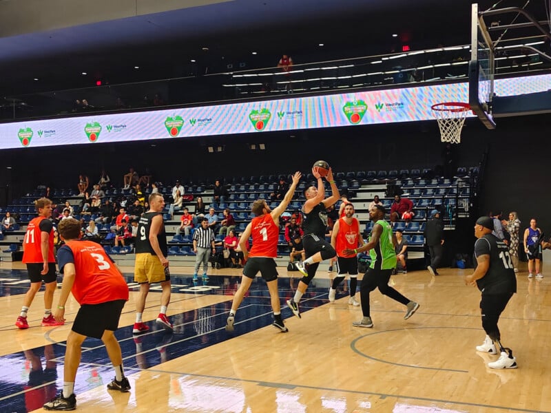 Basketball game in progress at an indoor court. Players in red jerseys face off against players in black and green jerseys. A player in black leaps to shoot the ball while others attempt to block. Spectators watch from the stands.
