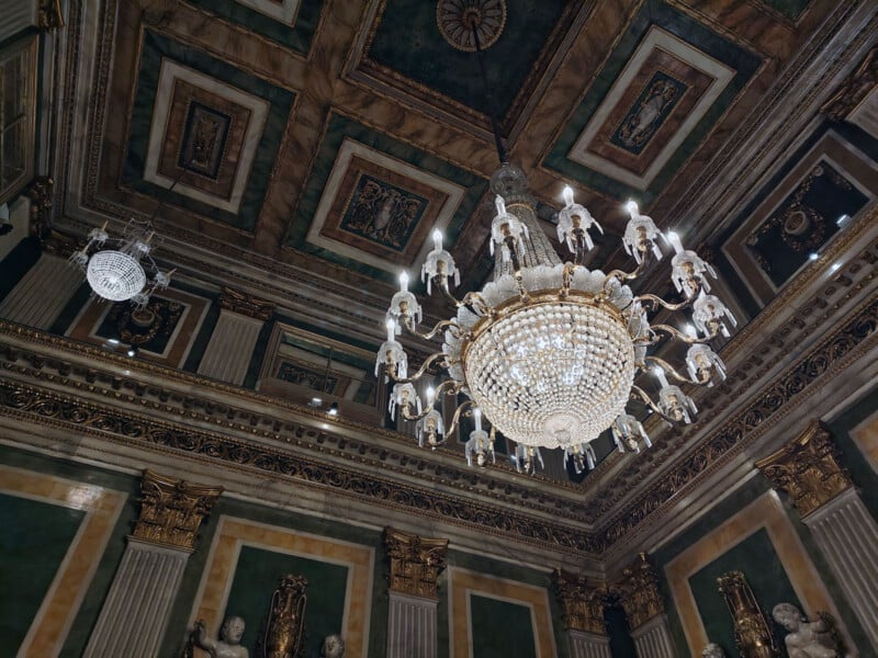 A grand interior with ornate chandeliers hangs from an intricately decorated ceiling. The room features luxurious green and gold accents with classical architectural elements, including statues and elaborate moldings.