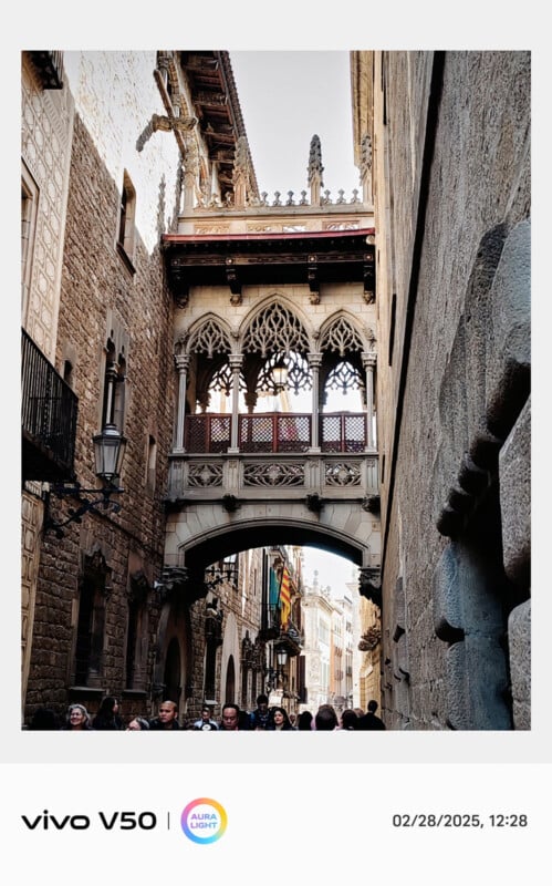 Ornate Gothic bridge connecting two stone buildings over a narrow street. The bridge has intricate carvings and arches. People walk beneath it, and sunlight illuminates the passage. "Vivo V50" and date are displayed on the border.