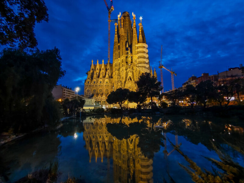 A nighttime scene of the Sagrada Familia in Barcelona, beautifully illuminated. The basilica's intricate architecture is reflected in a calm pond. Dark trees frame the foreground, and cranes are visible against a deep blue sky.