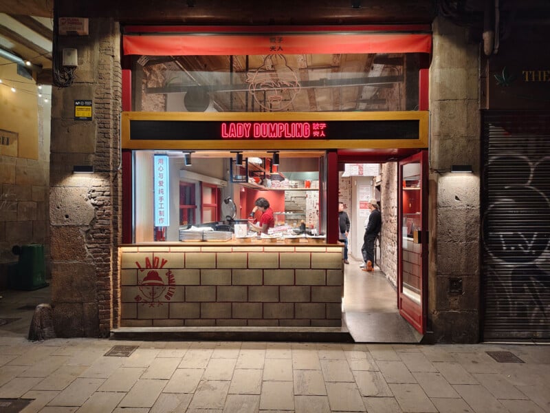 Street view of "Lady Dumpling" restaurant with red signage and an open serving counter. Inside, a person in a red shirt prepares food, while another customer stands nearby. The warm light from within contrasts with the dimly lit stone pavement outside.