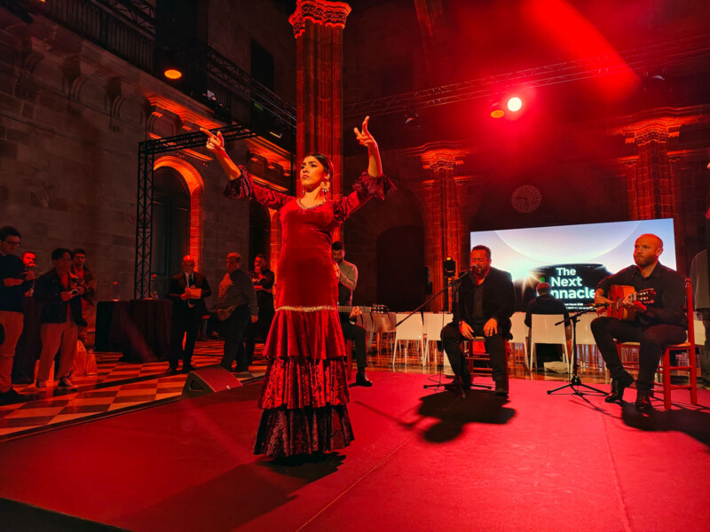 A flamenco dancer in a red dress performs on stage under red lighting. She is surrounded by musicians playing guitars. A brick wall and arches are visible in the background, along with a screen displaying "The Next Monadic." An audience watches the performance.