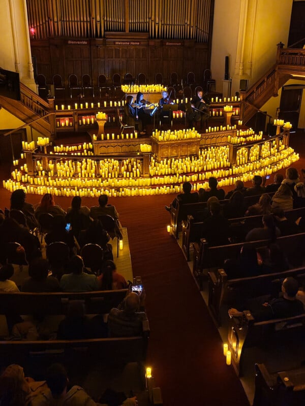 A dimly lit concert in a church features musicians on stage surrounded by numerous glowing candles. The audience sits in pews, watching the performance. Ornate wooden architecture and organ pipes are visible in the background.