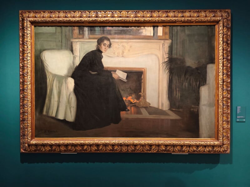 A framed painting of a woman in a black dress sitting in a white armchair by a fireplace. She holds a book and gazes towards the viewer. The scene is set within an ornate gold frame, hung on a teal wall.