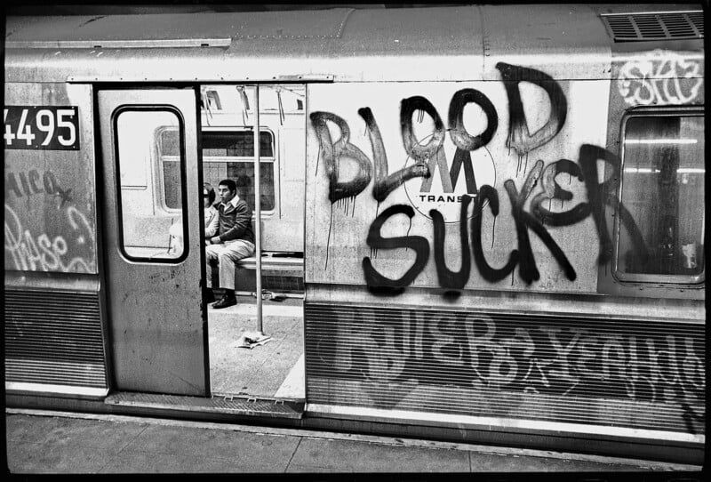 A man sits alone inside a subway car, visible through the open door. The subway’s exterior is covered in graffiti, with the words "BLOOD SUCKER" sprayed in large letters.