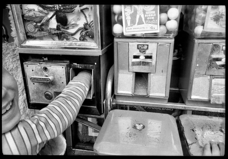 A child smiles while reaching into a toy vending machine, with bubble gum and trinket machines next to it. Only the child’s arm and part of their face are visible. The machines appear old and worn.