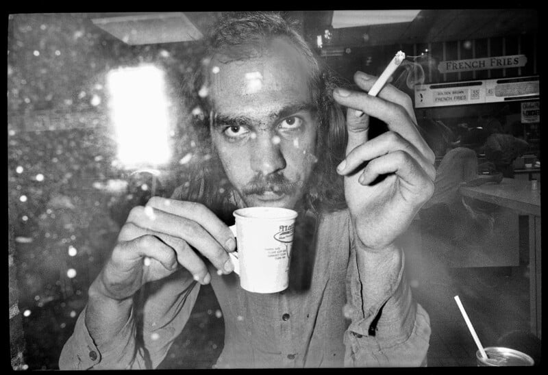 A man with long hair sits behind a dirty glass, holding a cup in one hand and a cigarette in the other, staring directly at the camera. The background includes a diner counter and a "French Fries" sign.