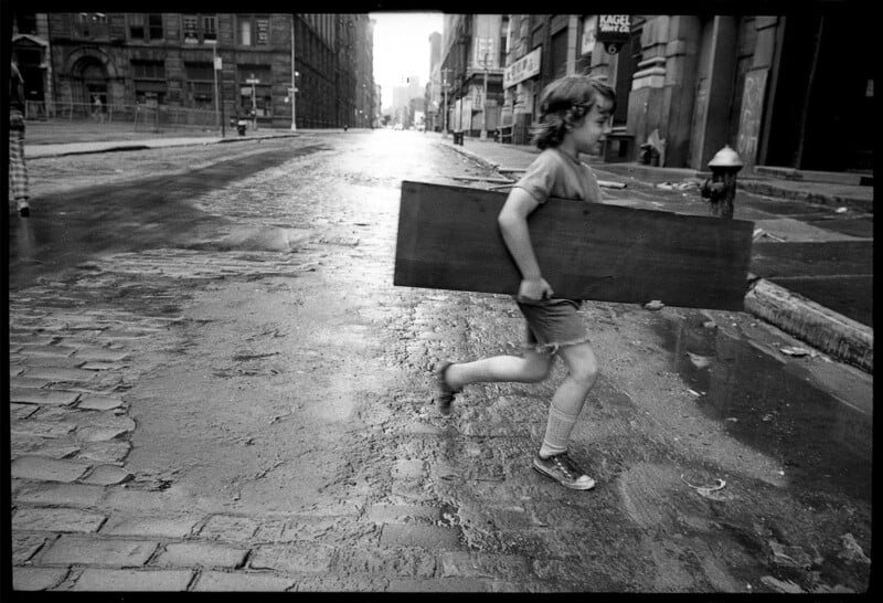 A young child in shorts and sneakers runs across an empty cobblestone street, carrying a large wooden board, with old buildings and a fire hydrant in the background.