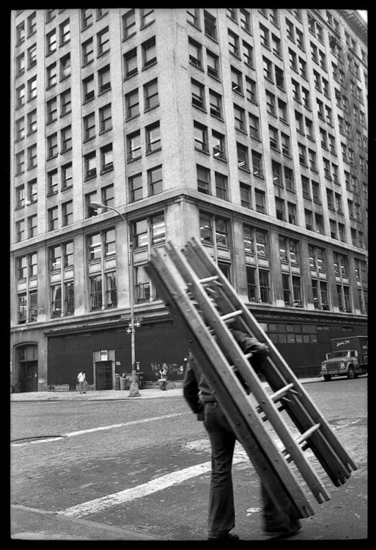 A person carrying a large wooden ladder walks across a city street, with a tall, multi-story building and a parked truck in the background. The photo is in black and white.