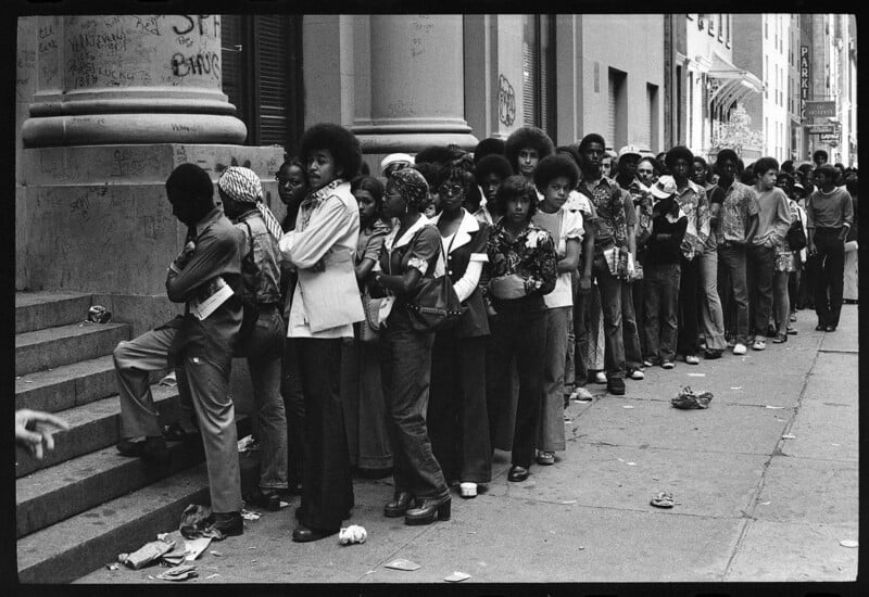 A long line of young people, mostly teenagers, stands on a city sidewalk near a building with graffiti-covered walls. Many wear 1970s-style clothing, including flared pants and afros. Some look at the camera, while others wait patiently.