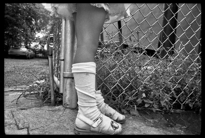 A black-and-white photo shows a child's legs in white socks and sandals, standing on tiptoes near a chain-link fence with overgrown plants and a car in the background.
