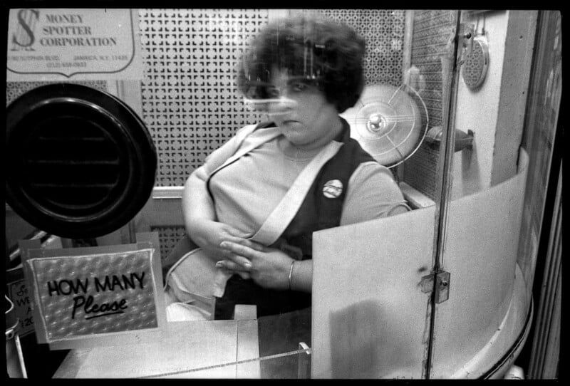 A woman with curly hair sits behind glass in a ticket booth, arms folded, wearing a vest and a button. A sign in front reads "HOW MANY Please." A fan and office items are visible behind her.