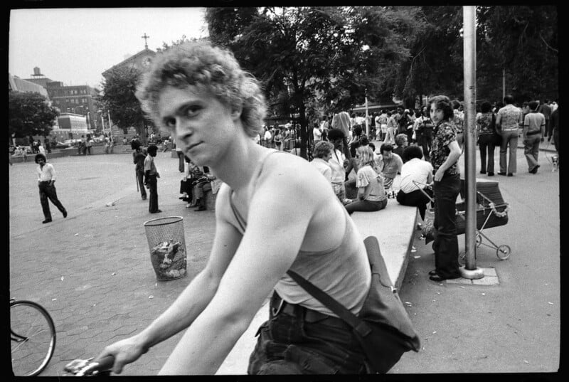 A young man with curly hair and a serious expression rides a bicycle in a busy park. Several people are sitting and standing in the background, and trees and buildings are visible. The image is in black and white.