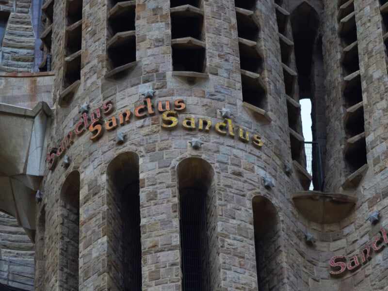 Detail of a stone building with multiple narrow, vertical windows. The word "Sanctus" is prominently displayed in stylized lettering across the arches. The architecture features rough-hewn stone and intricate designs.