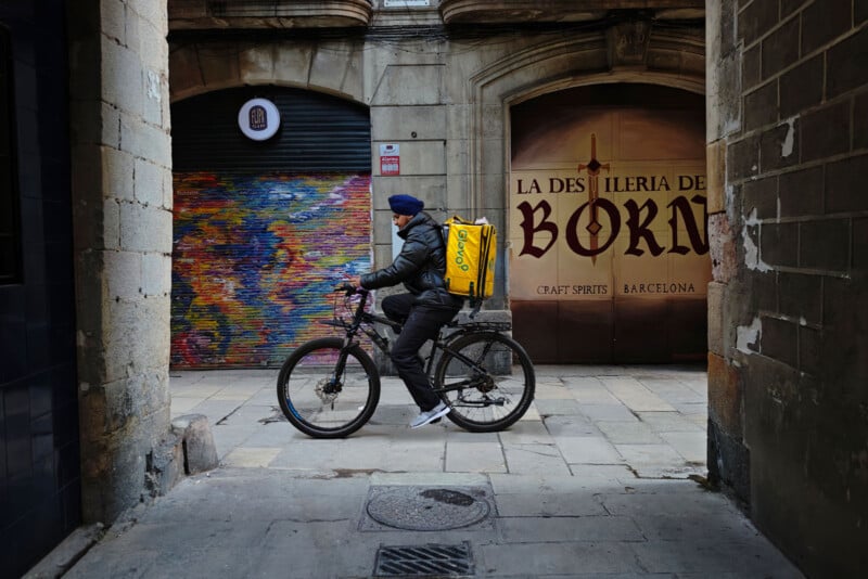 A person in a black jacket and helmet rides a bicycle with a yellow delivery bag on a narrow street. The background features colorful graffiti and a sign for "La Destileria de Born" in Barcelona.