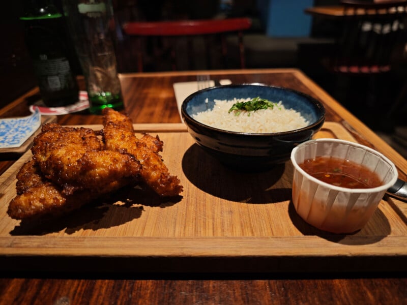 A wooden tray holds a bowl of white rice garnished with herbs, a serving of breaded fried chicken strips, and a small plastic cup of dipping sauce, all placed on a wooden table in a restaurant setting.