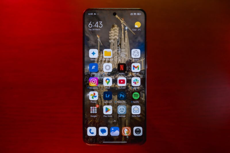 A smartphone displaying a variety of app icons on its screen, including a weather forecast showing 7°C and clear skies. The background image features a night view of a lit-up cathedral. The phone is set against a dark red background.