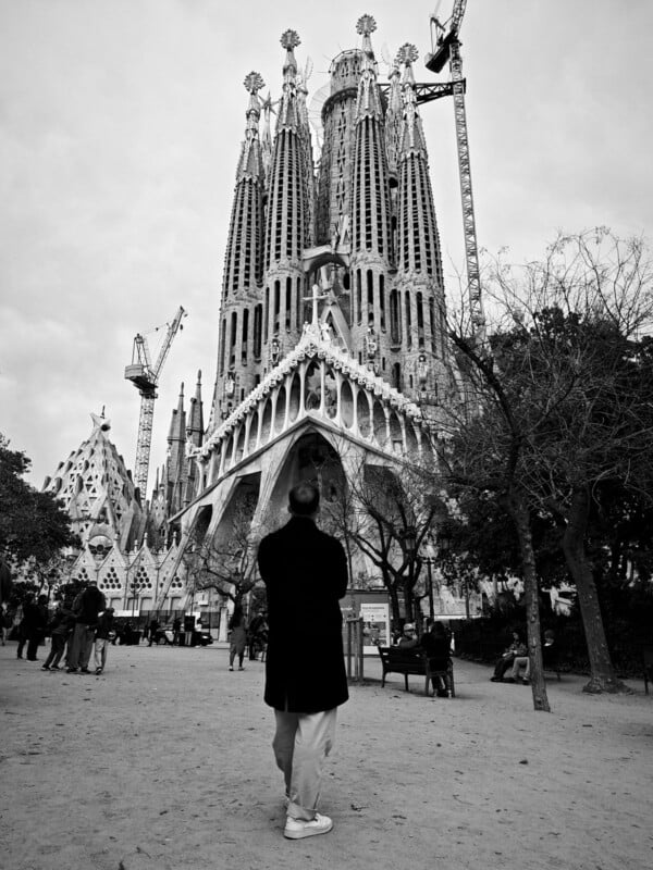A person in a long coat stands in front of the Sagrada Familia, a large, unfinished basilica in Barcelona. The structure features intricate spires and cranes are visible, indicating ongoing construction. The sky is overcast.