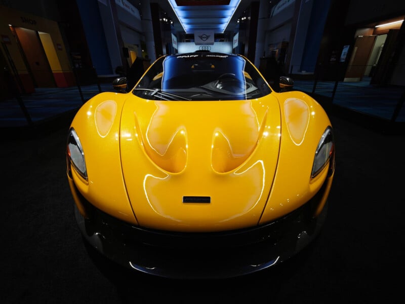 A sleek yellow sports car is displayed indoors under bright lighting. The car has a shiny, aerodynamic design with prominent curves, a black front splitter, and a visible logo on the hood. The setting conveys an upscale, modern showroom ambiance.