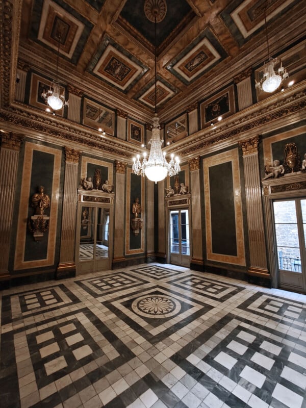 Ornate room with intricate geometric marble flooring, high ceilings with chandeliers, gold-accented walls, and decorative sculptures. Reflections are visible in large mirrored panels on the walls. Tall windows overlook a view outside.