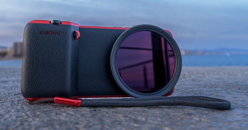 A camera with a red and black design rests on a concrete surface. It features a circular lens filter and a wrist strap. The background shows a blurred seascape with hints of a distant shoreline under a cloudy sky.