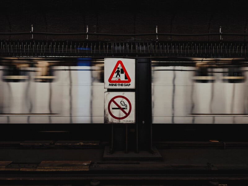 A subway station with a fast-moving train in the background. In the foreground, there are two signs: one warns to "Mind the Gap" and the other indicates no smoking. The image captures the motion blur of the train.
