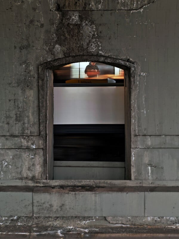 View through a rectangular window in a weathered concrete wall. A person wearing an orange shirt and mask is visible outside, blurred by motion. The scene suggests a moving train or subway, with a structured urban environment.