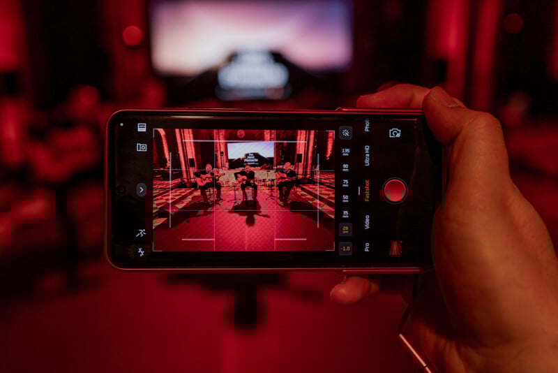 A person holds a smartphone capturing a red-tinted photo of a live band performing in a dimly lit venue. The screen displays the band and stage under red lighting. In the background, a blurred projection screen and audience are visible.