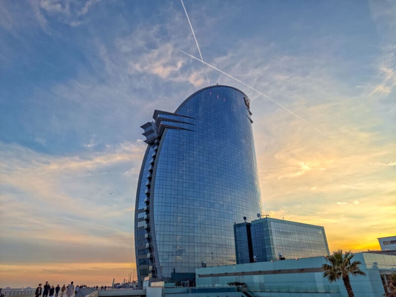 A modern, sail-shaped skyscraper with a glass exterior reflecting the sunset sky. A palm tree stands nearby, and a few people are visible on the ground. Vapor trails cross in the sky above.